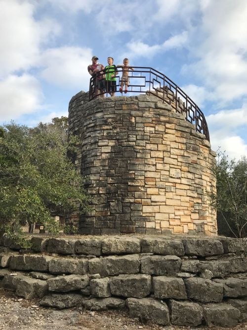 Three people stand on a stone tower with a railing, surrounded by trees and a cloudy sky.