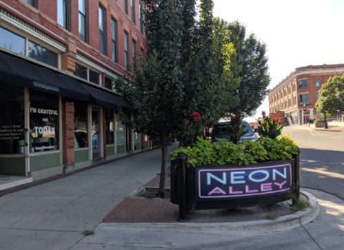 A street view featuring a sign that reads "NEON ALLEY" surrounded by greenery and flowers in a vibrant urban setting.