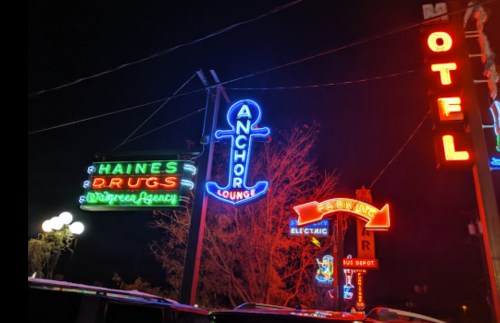 Colorful neon signs for Haines Drugs, Anchor Lounge, and a parking area, illuminated against a night sky.