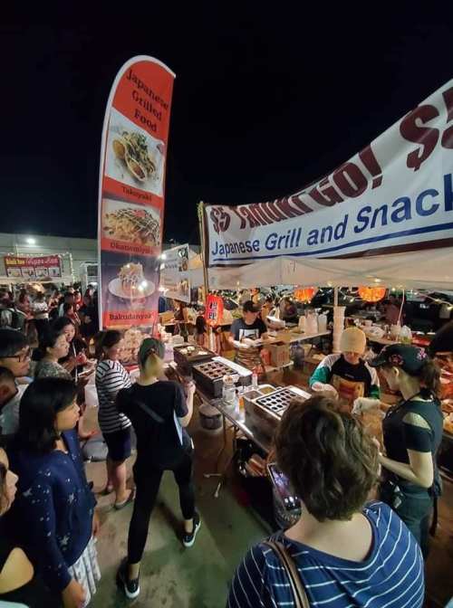 A bustling night market scene with a Japanese grill stall, featuring a crowd enjoying food and vibrant banners.
