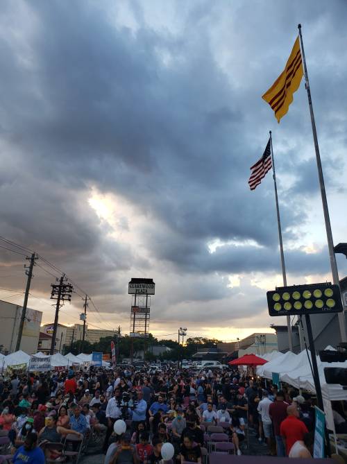 A large crowd gathers at an outdoor event under cloudy skies, with American and Vietnamese flags flying above.