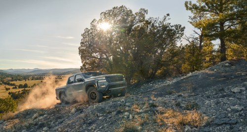 A rugged truck driving on a rocky terrain with dust rising, surrounded by trees and a sunset in the background.