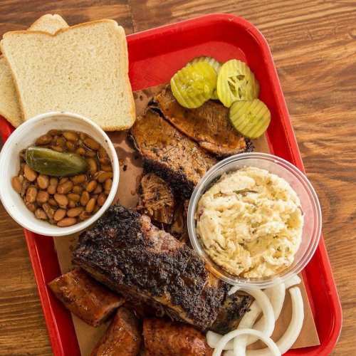 A red tray with sliced meats, pickles, bread, beans, and onion slices on a wooden table.