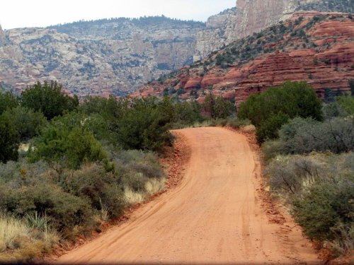 A winding dirt road surrounded by red rock formations and sparse vegetation in a rugged landscape.