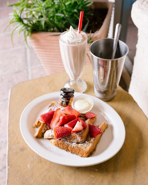 A plate of French toast topped with strawberries and powdered sugar, alongside a milkshake and a metal cup.
