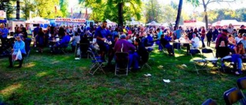 A bustling outdoor festival scene with crowds of people sitting at tables and food stalls in the background.