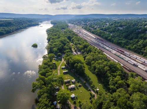 Aerial view of a river, green trees, and a train yard, with a campground visible along the riverbank.