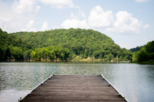 A wooden dock extends into a calm lake, surrounded by lush green hills and a blue sky with fluffy clouds.