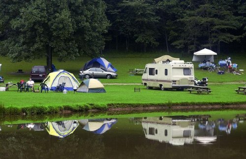 A serene campsite by a lake, featuring tents, an RV, and people enjoying the outdoors amidst lush greenery.