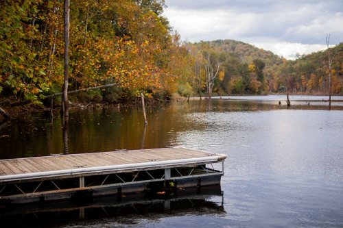 A wooden dock extends over a calm lake surrounded by autumn foliage and distant hills under a cloudy sky.