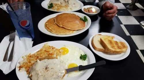 A table set with plates of breakfast: pancakes, eggs, hash browns, toast, and a drink.