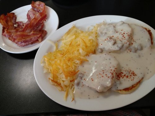 A plate of biscuits and gravy with hash browns, accompanied by a side of crispy bacon.