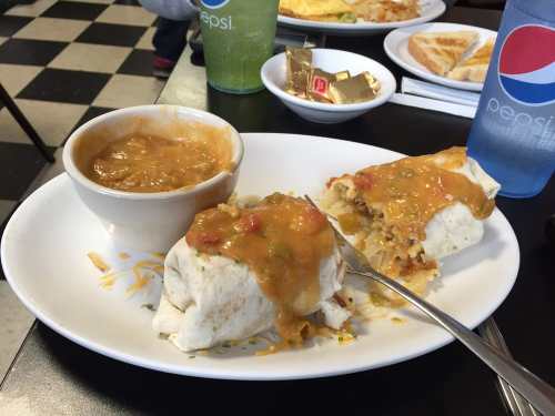 A plate with two burritos topped with sauce, a bowl of soup, and drinks in a diner setting.