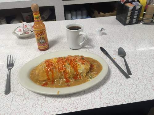 A plate of enchiladas topped with sauce and cheese, alongside a cup of coffee and hot sauce on a diner table.