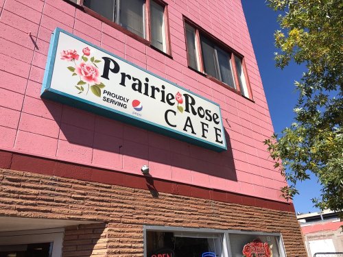 Sign for Prairie Rose Café, featuring floral designs and the text "Proudly Serving" on a pink building.