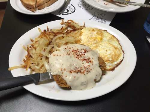 A plate with crispy hash browns, a fried egg, and chicken smothered in creamy gravy, served with toast in the background.