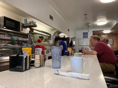 A diner interior with a counter, condiments, and two customers seated, one reading a menu and the other at the register.