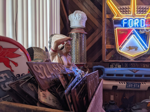 A collection of vintage license plates, a statue of a Native American, and a neon Ford sign in a rustic setting.