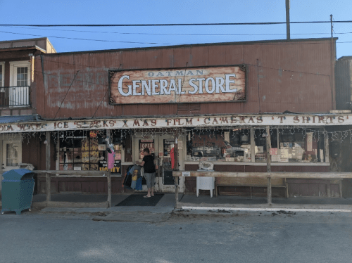 A rustic general store with a sign reading "Oatman General Store," featuring various items in the windows and a welcoming entrance.