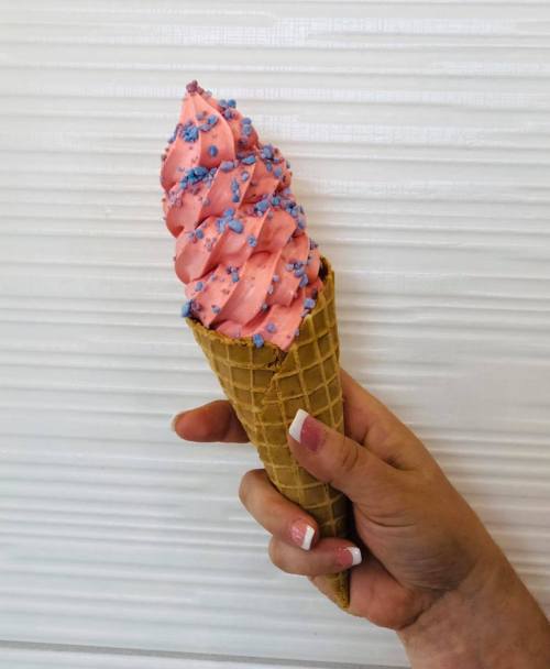 A hand holding a waffle cone filled with pink soft serve ice cream topped with blue sprinkles.