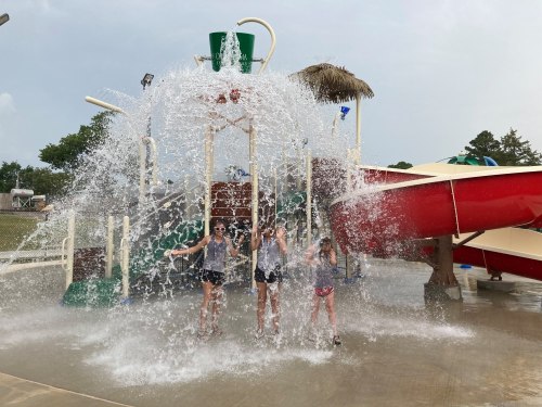 Three children play under a large water spray feature at a water park, with a red slide in the background.