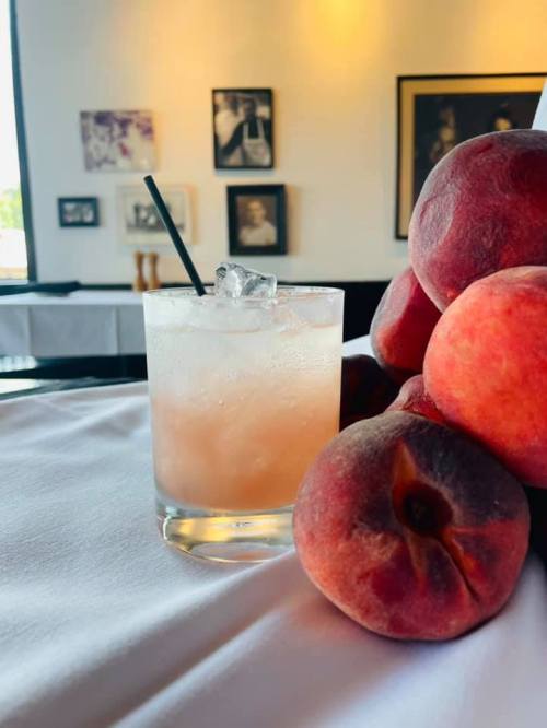 A refreshing cocktail with ice sits beside fresh peaches on a table, with framed photos in the background.
