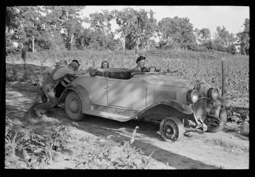A vintage car stuck in a field, with three people pushing it while another sits inside. Trees in the background.