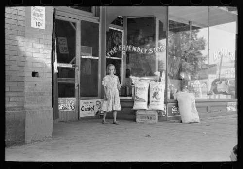 A young girl stands outside a store, surrounded by bags and vintage advertisements, in a black-and-white photo.