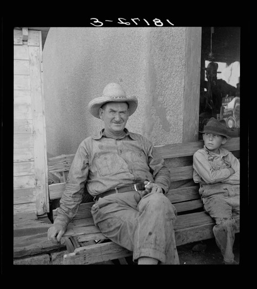 A man in a straw hat sits on a bench beside a boy, both looking at the camera, in a rustic outdoor setting.
