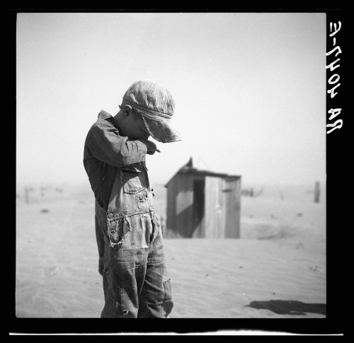 A young boy in overalls and a cap stands in a dusty landscape, looking down, with a small shed in the background.