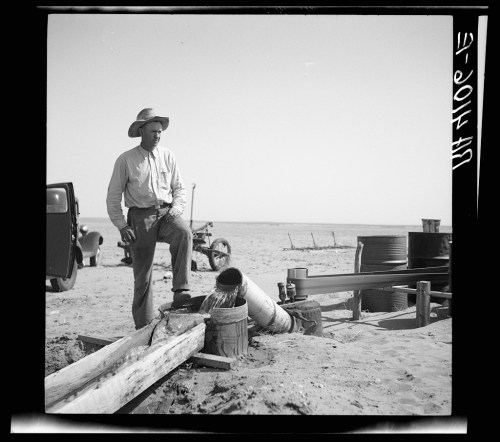 A man in a cowboy hat stands by a water pump in a desert landscape, with vintage vehicles in the background.