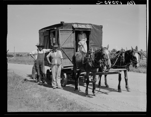 Two men stand beside a horse-drawn wagon on a dirt road, with one man wearing overalls and a hat.