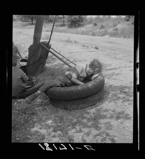A child rests on a tire in a rural setting, with another child nearby, surrounded by dirt and sparse vegetation.