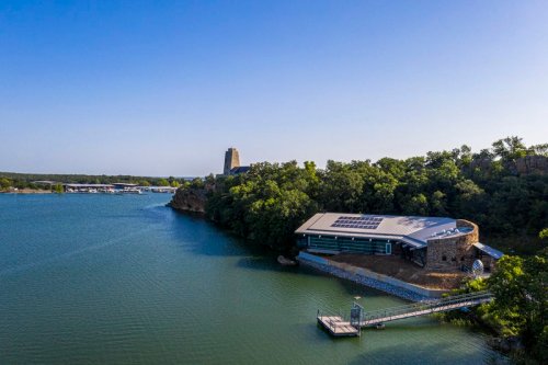 Aerial view of a modern building by a lake, surrounded by trees and a dock, with a tower in the background.
