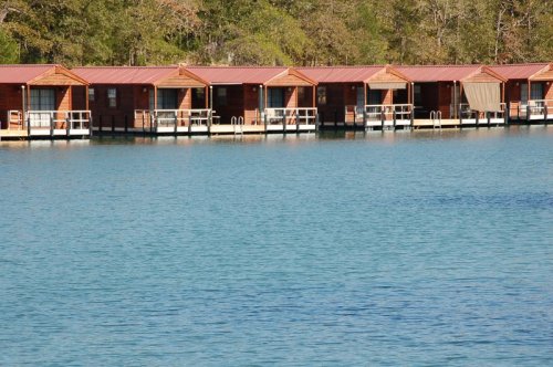 Row of wooden cabins with red roofs on a calm blue lake, surrounded by trees.