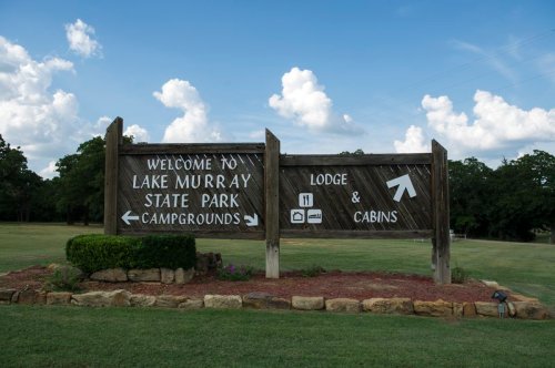 Welcome sign for Lake Murray State Park, featuring directions to campgrounds, lodge, and cabins against a blue sky.