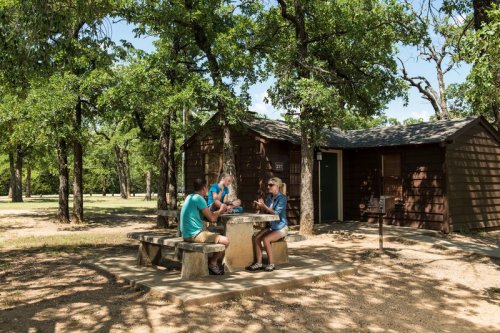 Two people sit at a picnic table near a cabin, surrounded by trees on a sunny day.