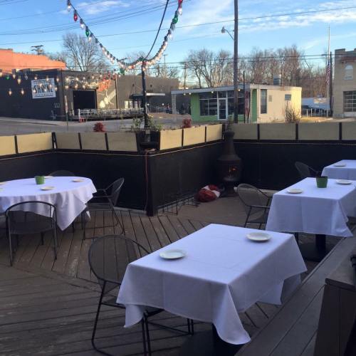 Outdoor dining area with empty tables set with white tablecloths, surrounded by a fence and string lights in the background.