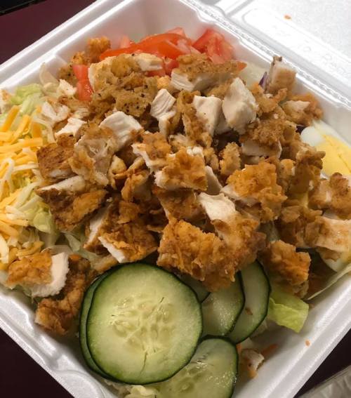 A salad with chopped fried chicken, lettuce, tomatoes, cheese, and cucumber slices in a takeout container.