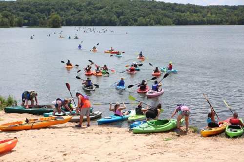 A busy lake scene with people kayaking and preparing kayaks on the sandy shore under a clear blue sky.