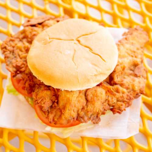 A crispy fried chicken sandwich with lettuce and tomato, served on a soft bun, on a yellow table.