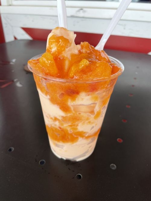 A cup of orange mango ice cream topped with a vibrant orange sauce, served with two plastic spoons.