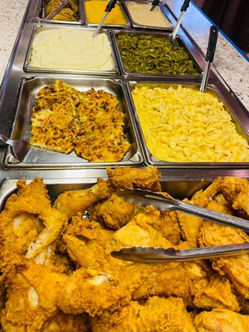 A buffet spread featuring fried chicken, mashed potatoes, green beans, pasta, and various sauces in metal trays.