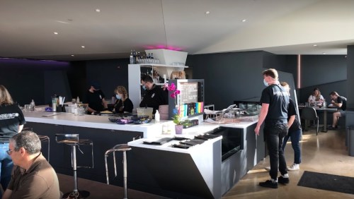 A modern café interior with staff preparing food, customers seated, and a bar area in a bright, open space.