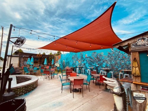 Outdoor seating area with colorful murals, shaded by a large canopy, and string lights overhead.
