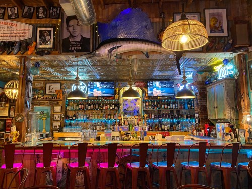 A cozy bar with a wooden counter, colorful lights, and shelves filled with bottles, featuring TVs and vintage decor.