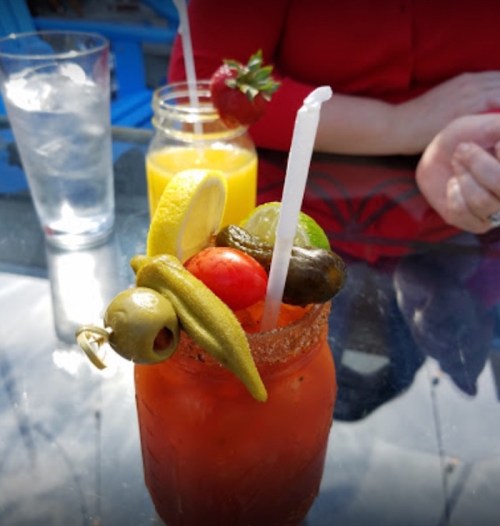 A close-up of a colorful cocktail garnished with olives, pickles, and a lemon slice, with drinks in the background.