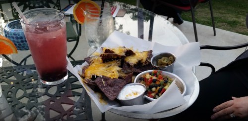A plate of nachos topped with cheese, served with dips and a colorful side salad, alongside two drinks on a table.