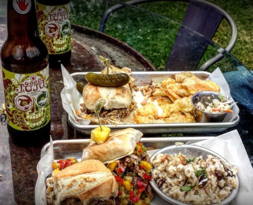 Two trays of food featuring sandwiches, shrimp, rice, and coleslaw, with two bottles of beer on a table outdoors.