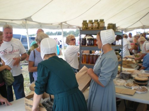 A busy outdoor market scene with people interacting, tables of food, and vendors in traditional attire under a tent.